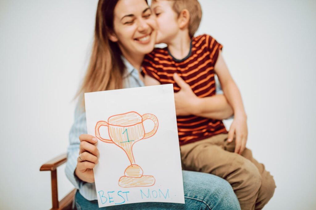 A cheerful mother receives a hug and a drawing labeled 'Best Mom' from her young son.